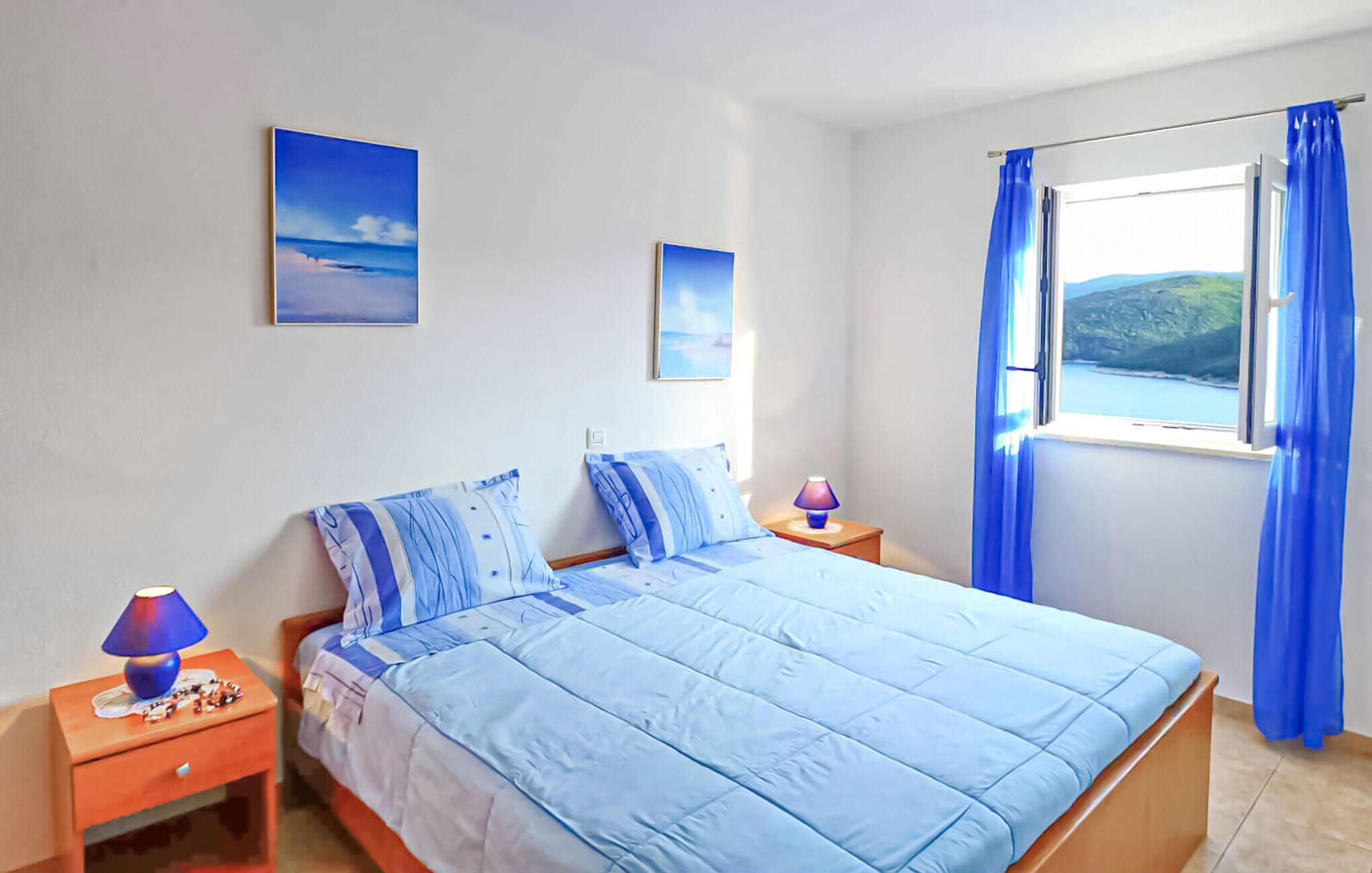 A bedroom with a bed, two pillows, a nightstand with a lamp, and a window with curtains.