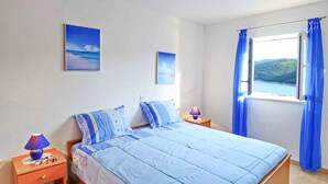 A bedroom with a bed, two pillows, a nightstand with a lamp, and a window with curtains. - 4 bedroom beautiful home in Rabac (Rabac)