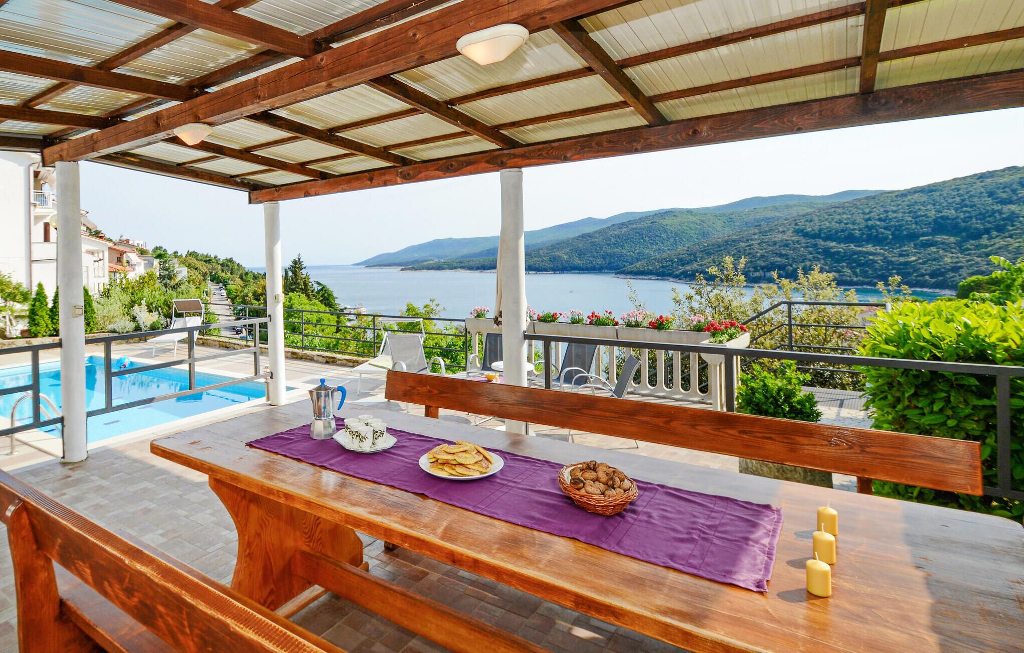 A wooden table with a purple tablecloth, a white pitcher, a plate of food, a basket of bread, and candles on a terrace overlooking a pool and a scenic view of mountains and water.