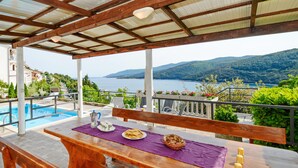 A wooden table with a purple tablecloth, a white pitcher, a plate of food, a basket of bread, and candles on a terrace overlooking a pool and a scenic view of mountains and water. - 4 bedroom beautiful home in Rabac (Rabac)