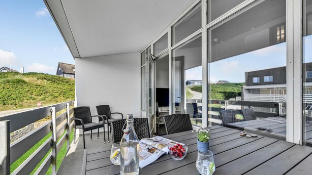 A covered outdoor area with a wooden deck, wicker chairs, a table with a magazine, a glass bottle, and a bowl of fruit. - Gorgeous apartment in Ringkøbing (Ringkøbing)