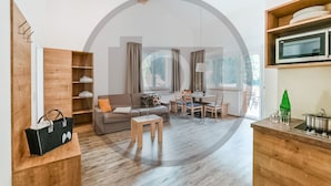 Uma sala de estar moderna com sofá, mesa de centro e área de jantar. Há uma estante de madeira e uma cozinha com micro-ondas e uma garrafa verde.