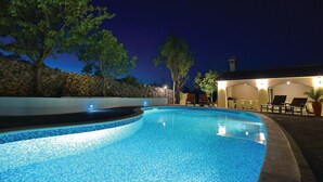 Una piscina con muro de piedra y árboles, rodeada por un patio con mobiliario exterior y un edificio iluminado.