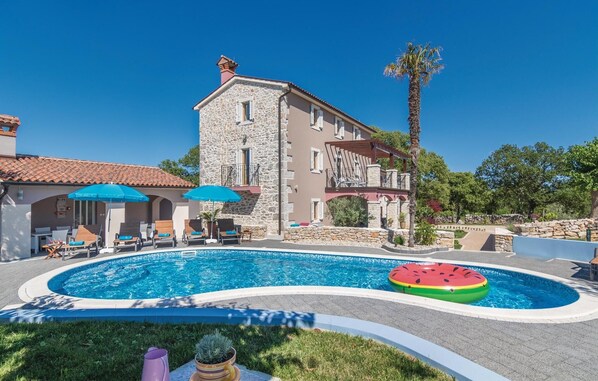 Zona piscina con lettini e ombrelloni, un edificio in pietra con balcone e una palma.