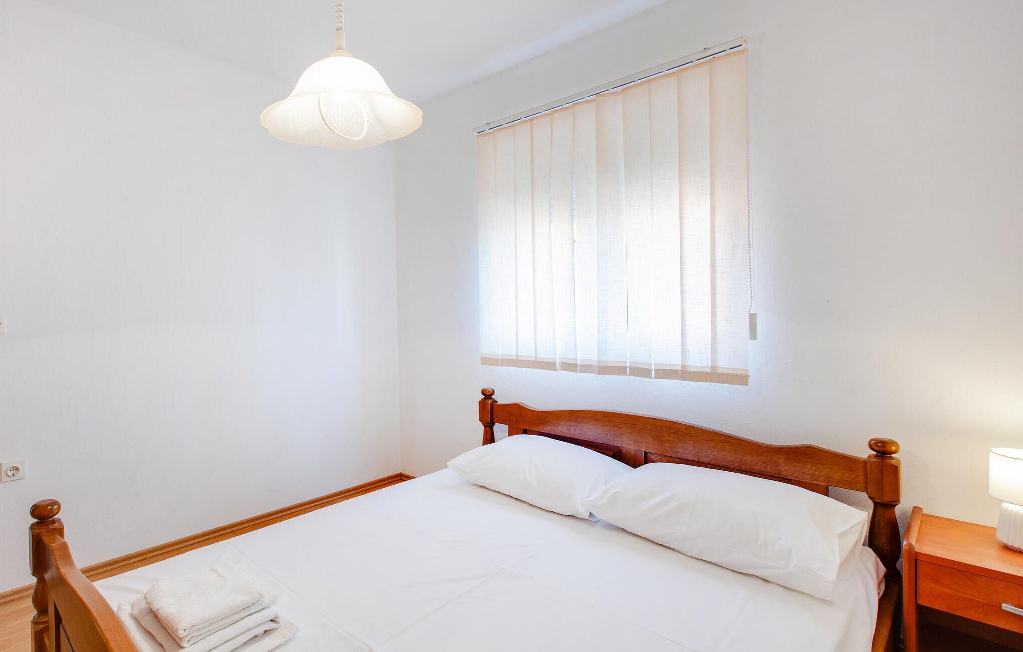 A neatly made bed with white linens and pillows, a wooden headboard, a bedside table with a lamp, and a window with vertical blinds.