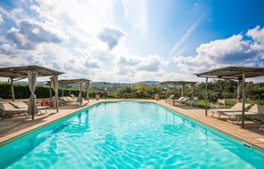 Ein klares Schwimmbecken mit Sonnenschirmen und Liegestühlen, umgeben von einem angelegten Garten und Blick auf ferne Hügel.
