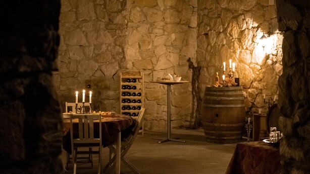 A dimly lit cellar with stone walls, a wooden table with candles, a wine rack, and a barrel.