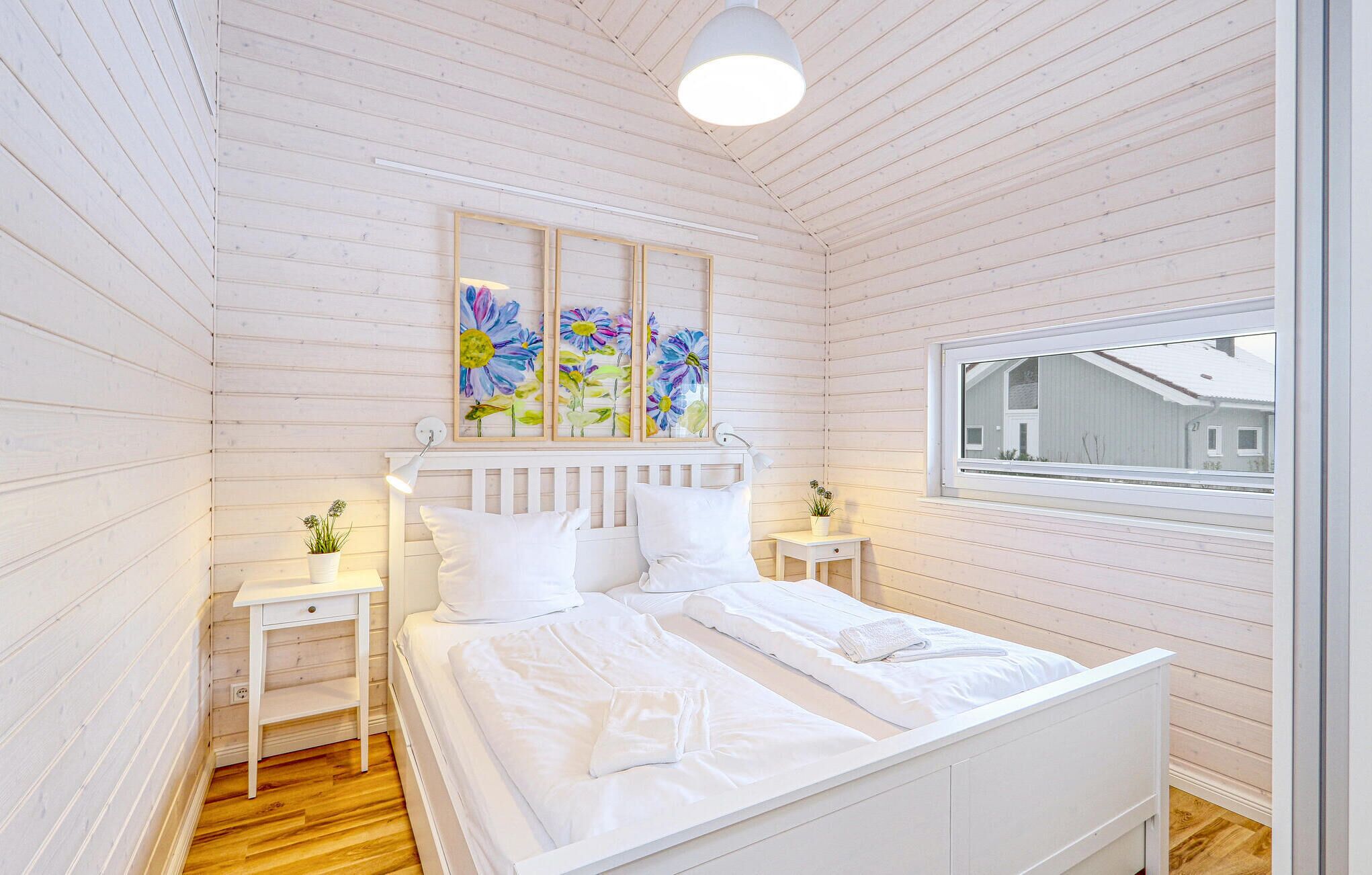 A bedroom with a wooden slat ceiling, a bed with white bedding, two bedside tables with lamps, and a window with a view of a house.