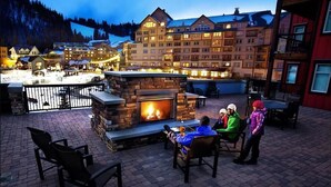 An outdoor patio with a stone fireplace, seating, and a view of a large hotel building in the background. - Location and Comfort - One Bedroom Ski In / Ski out, Hot Tub and Fire Pit (Winter Park)