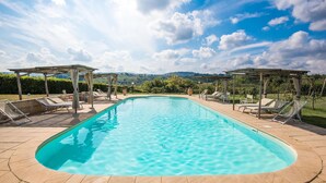 A rectangular swimming pool with clear blue water, surrounded by lounge chairs and wooden pergolas under a partly cloudy sky. - 2 bedroom cozy home in Montaione (Montaione)