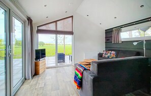 Ein modernes Wohnzimmer mit einer Couch, einem Fernseher und Blick auf eine grüne Landschaft durch große Fenster.