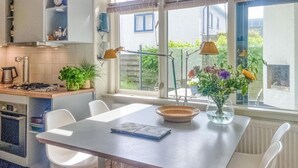 Ett modernt kök med vita skåp, ett matbord med stolar, en vas med blommor och ett fönster med utsikt över omgivningen.
