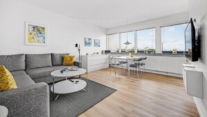 Ein modernes Wohnzimmer mit einem grauen Sofa, einem Couchtisch und einem Essbereich mit weißem Tisch und Stühlen. Große Fenster bieten Blick auf Himmel und Grünflächen.