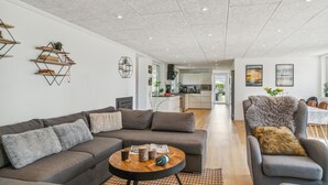 Un salón moderno con un sofá gris, una mesa de centro redonda y un sillón gris con un plaid de piel. Se observan estanterías de madera en la pared y una cocina al fondo.