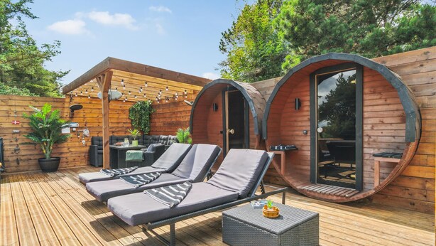Dos cabañas de madera con puertas correderas, una terraza de madera, sillones de descanso y una pequeña mesa con una planta.