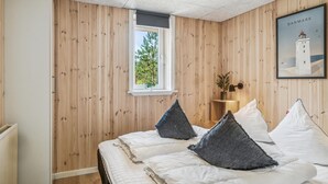 Un dormitorio con paredes de madera panelada, una ventana con vistas a árboles, una cama con ropa blanca y dos almohadas oscuras, una mesita de noche con una lámpara y un cuadro con una imagen de un faro.