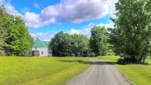 Un chemin de campagne mène à une maison blanche dotée d’un toit vert, entourée d’une végétation luxuriante et d’arbres.