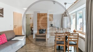 Ein modernes Wohnzimmer mit einer Couch, einem Esstisch mit Stühlen, einer Küchenzeile und einem großen Fenster mit Vorhängen.