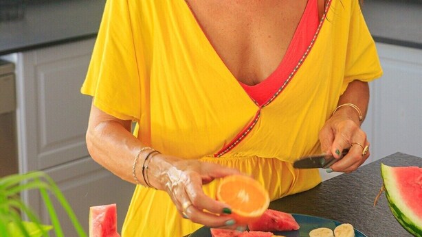 Una persona con un vestito giallo taglia anguria e arance in una cucina con un forno a microonde e degli armadietti.