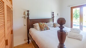 Una camera da letto con una testiera in legno, due letti con lenzuola bianche, cuscini decorati, lampade da comodino e vista sull'esterno tramite una porta.