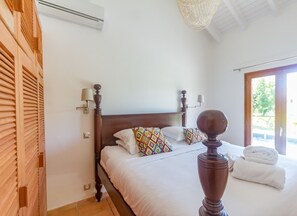 Un dormitorio con cabecera de madera, dos camas con ropa de cama blanca, almohadas estampadas, lámparas de noche y vistas al exterior a través de una puerta.