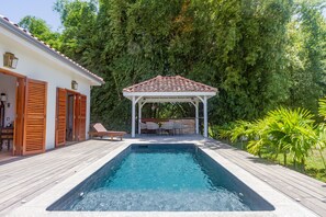 A swimming pool with a wooden deck, a covered seating area, and lush greenery in the background.