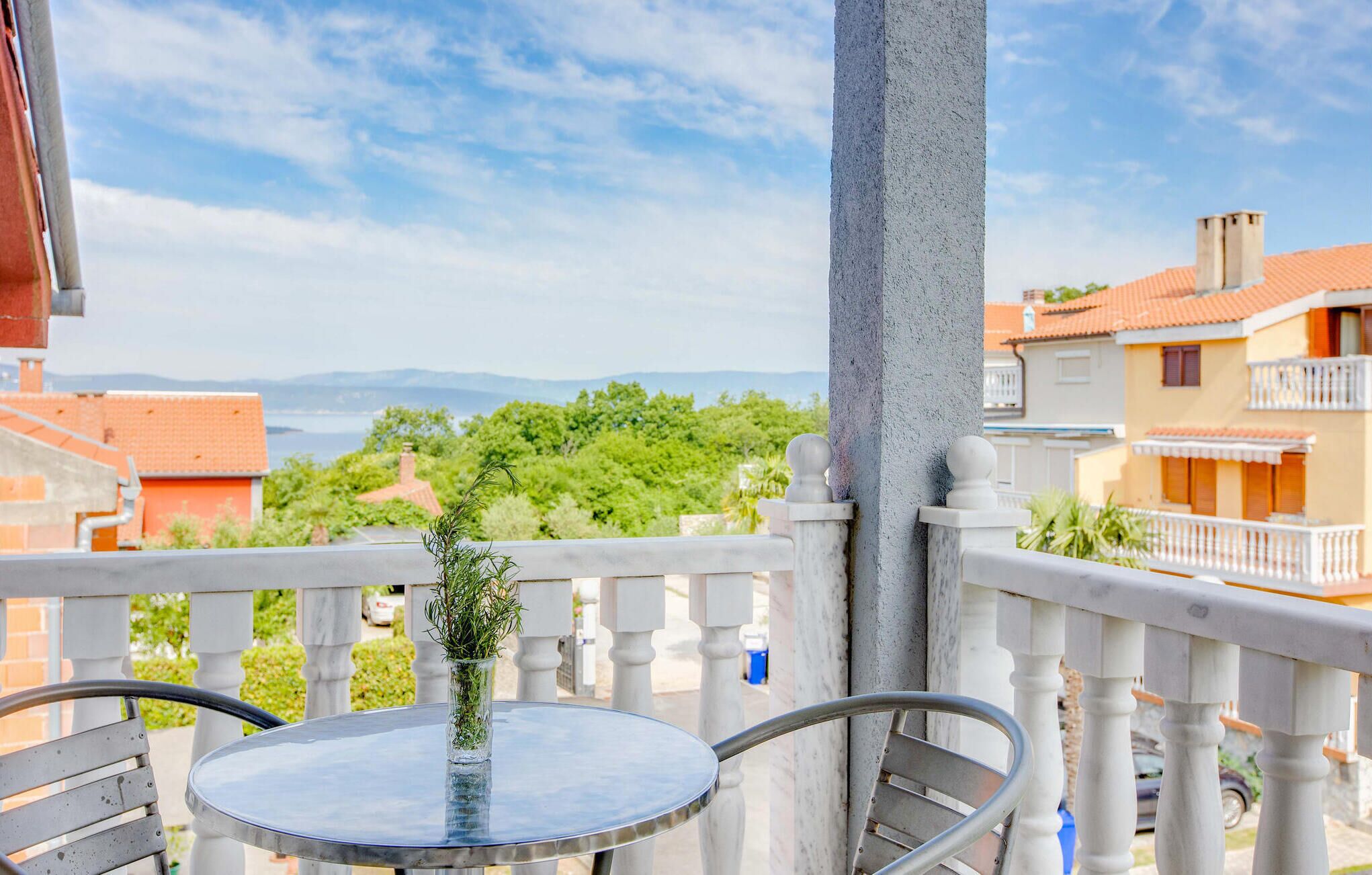 En balkon med et rundt bord og stole, en vase med grønne planter, og udsigt til bygninger og fjerne bjerge.