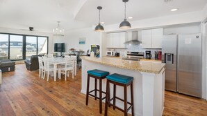 A modern kitchen with a central island, stainless steel appliances, and a dining area with white chairs. - Beach Paradise - Perfect Ocean Sunset Views with Pool & Sauna & Breakfast! (Mexico Beach)