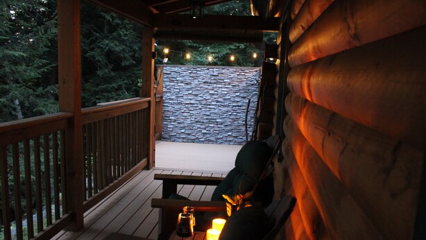 Eine Holzterrasse mit einer Steinsäule, einer Bank und einem Tisch mit Kerzen und einem Glas.
