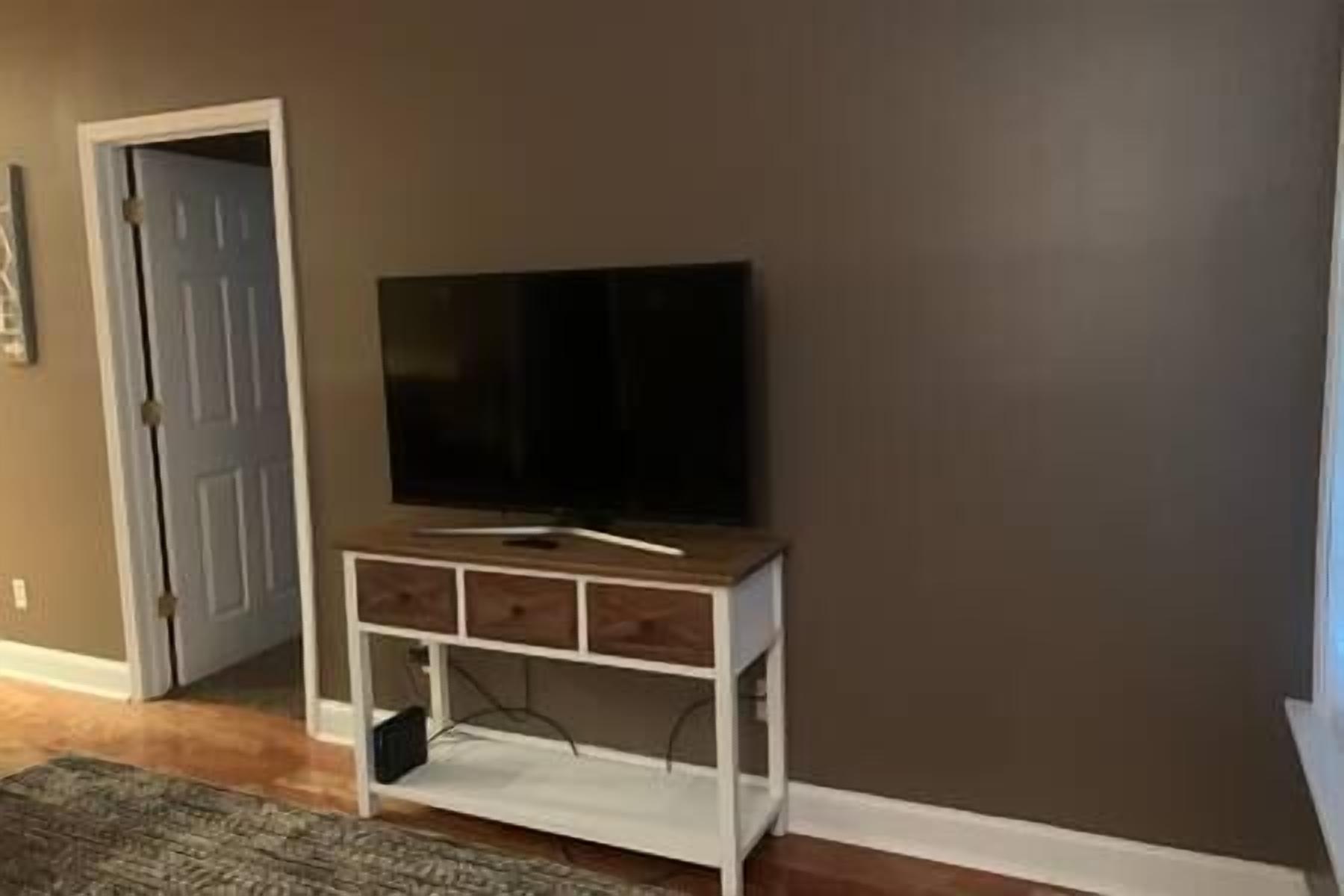 A room with a television on a wooden stand against a dark wall, a door to the left, and a patterned rug on the floor.