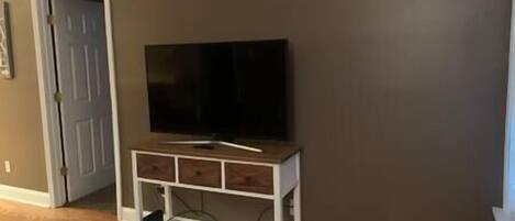 A room with a television on a wooden stand against a dark wall, a door to the left, and a patterned rug on the floor.