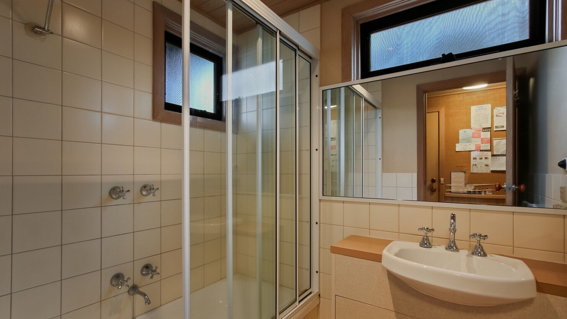 A modern bathroom with a glass shower enclosure, a sink with two faucets, and a mirror reflecting a door with a bulletin board.