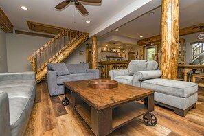 Un vaste salon avec un escalier en bois, des canapés gris, une table basse en bois et, au fond, un coin cuisine.