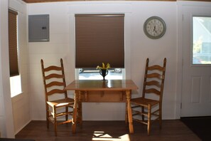 Een eetgedeelte met een houten tafel en twee stoelen, een raam met zonwering, een klok aan de muur en een kleine plant op de tafel.