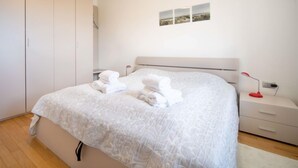 A neatly made bed with white linens and towels, a white wardrobe, a bedside table with a lamp, and a wall-mounted picture.