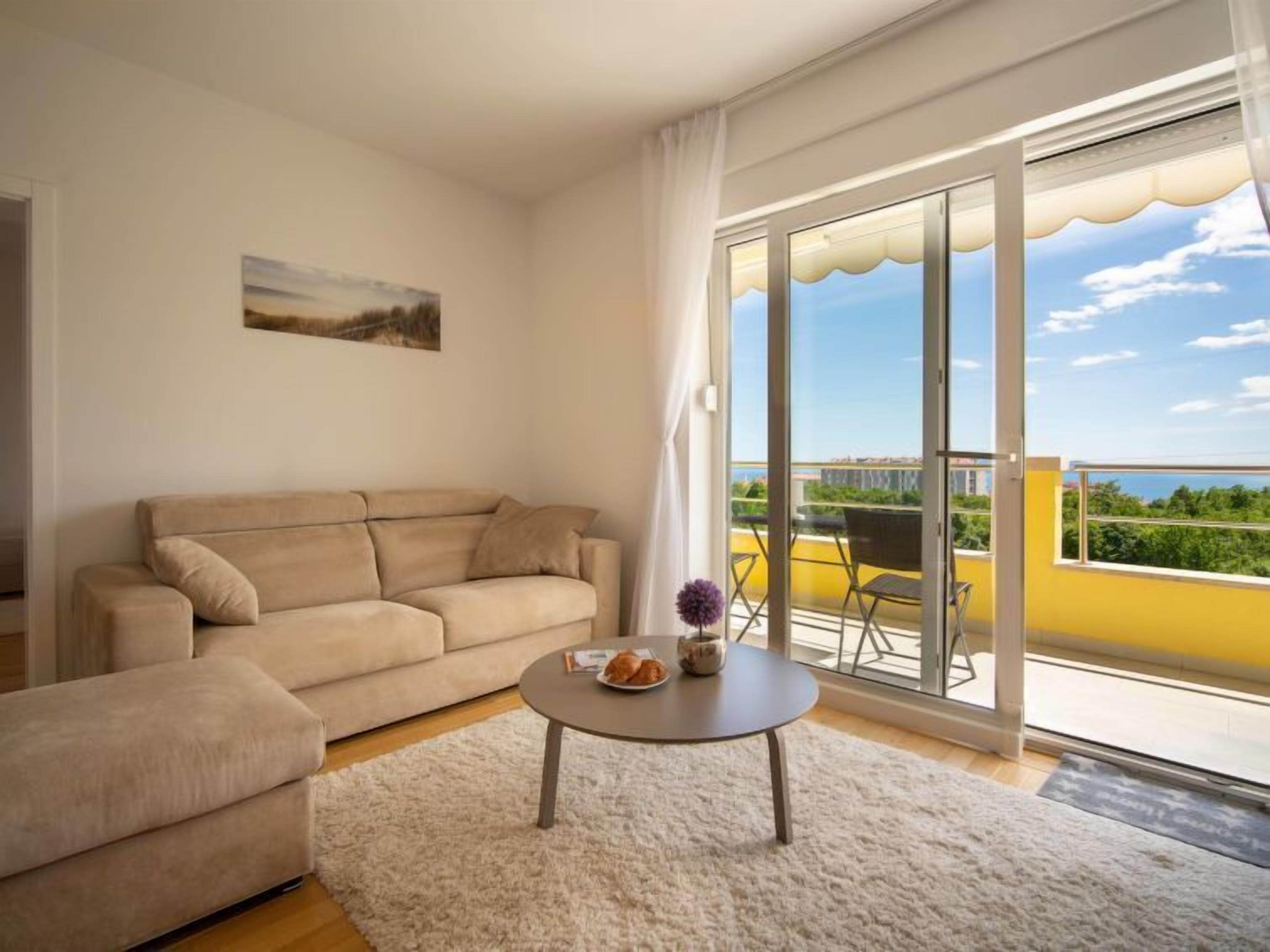 A living room with a beige sofa, a round coffee table, and a view of a balcony with outdoor furniture.