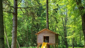 Ein hölzernes Spielhaus mit gelbter Rutsche in einem bewaldeten Gebiet.