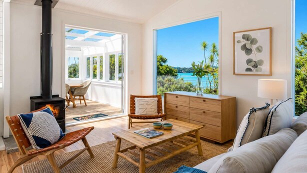 A cozy living room with a fireplace, a large window with a view of the sea, a wooden coffee table, and a comfortable sofa. - Bluebelle - Huruhi Bay Sea Views (Waiheke Island)