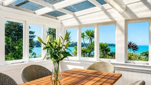A sunroom with a wooden table and wicker chairs, large windows with a view of the sea and greenery. - Bluebelle - Huruhi Bay Sea Views (Waiheke Island)