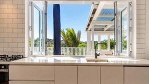 A modern kitchen with an open layout, a gas stove, and a view of a balcony with a white railing and greenery. - Bluebelle - Huruhi Bay Sea Views (Waiheke Island)