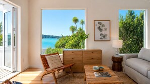 A room with a wooden coffee table, a sofa, a chair, a wooden cabinet, and a view of the sea. - Bluebelle - Huruhi Bay Sea Views (Waiheke Island)