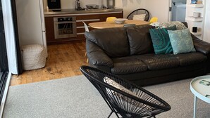 Een moderne woonkamer met een zwarte leren bank, een zwarte stoel en een kleine ronde tafel. Achtergrond toont een keuken met houten kastjes en een spoelbak.