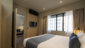 A hotel room with a large bed, a flat-screen TV mounted on the wall, a window with curtains, and a door leading to a sitting area. - Quest 256 Lambton (Wellington)