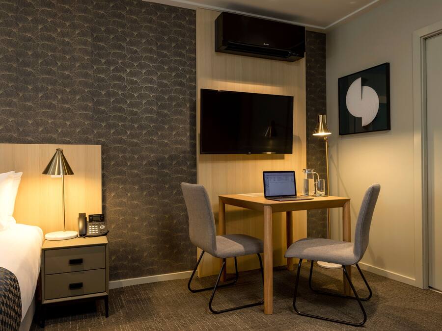 A hotel room with a bed, bedside table with a lamp, a desk with a laptop, two chairs, a flat-screen TV, and a painting on the wall.
