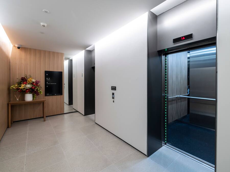 A modern hallway with an elevator, a reception desk with a floral arrangement, and a digital display.