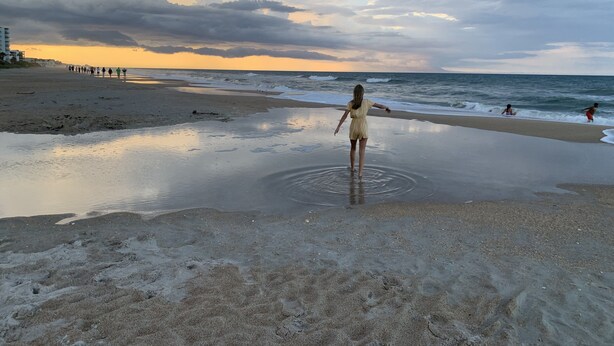 Una spiaggia con una persona che si trova in acque basse, un molo sullo sfondo e un cielo nuvoloso.