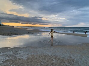 Ein Strand mit einer Person, die im flachen Wasser steht, einem Pier im Hintergrund und einem bewölkten Himmel.