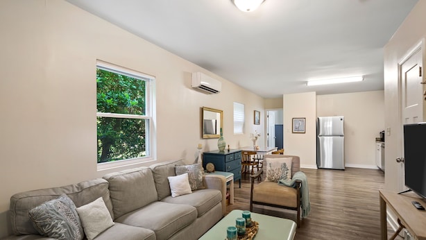 A modern living room with a sofa, coffee table, and dining area. There is a large window with a view of greenery. - Adorable Chesapeake Bay Beach Cottage B Just 1 Block from the Sand! (Virginia Beach)