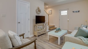 A living room with a TV on a wooden stand, a staircase, and a door. - Adorable Chesapeake Bay Beach Cottage B Just 1 Block from the Sand! (Virginia Beach)