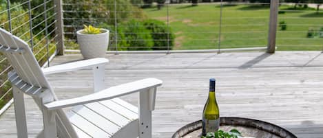 A wooden deck with a white chair, a rustic barrel, and a bottle of wine, overlooking a green landscape.